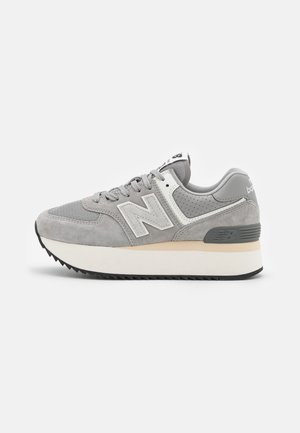 Grauer und weißer New Balance Sneaker mit Mesh- und Wildlederpaneelen, dicker weißer Sohle und schwarzer Profilsohle, seitlich auf weißem Hintergrund dargestellt.