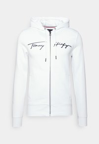 Sudadera blanca con cremallera, hecha de una tela suave, que presenta un logo negro, capucha con cordón y bolsillos laterales; textura lisa con puños acanalados.