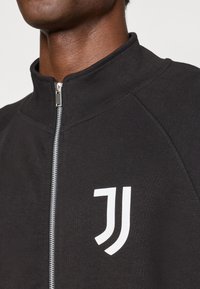 Crna zip-up jakna izrađena od mekog materijala, s visokim kragnom i bijelim logotipom sa lijeve strane. Prisutan je detalj šivanja.