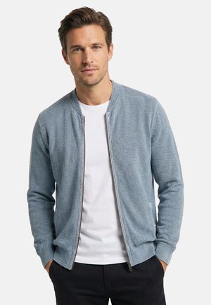 Mann trägt eine hellblaue Strickjacke mit Reißverschluss über einem weißen T-Shirt, Hände in den Taschen einer schwarzen Hose.