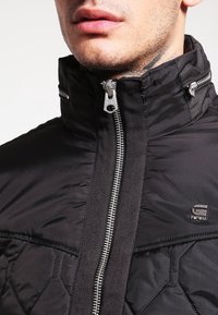 Svart vadderad jacka med quiltat mönster, hög krage och silverfärgad dragkedja. Logotypdetalj på vänster sida och slät textur.