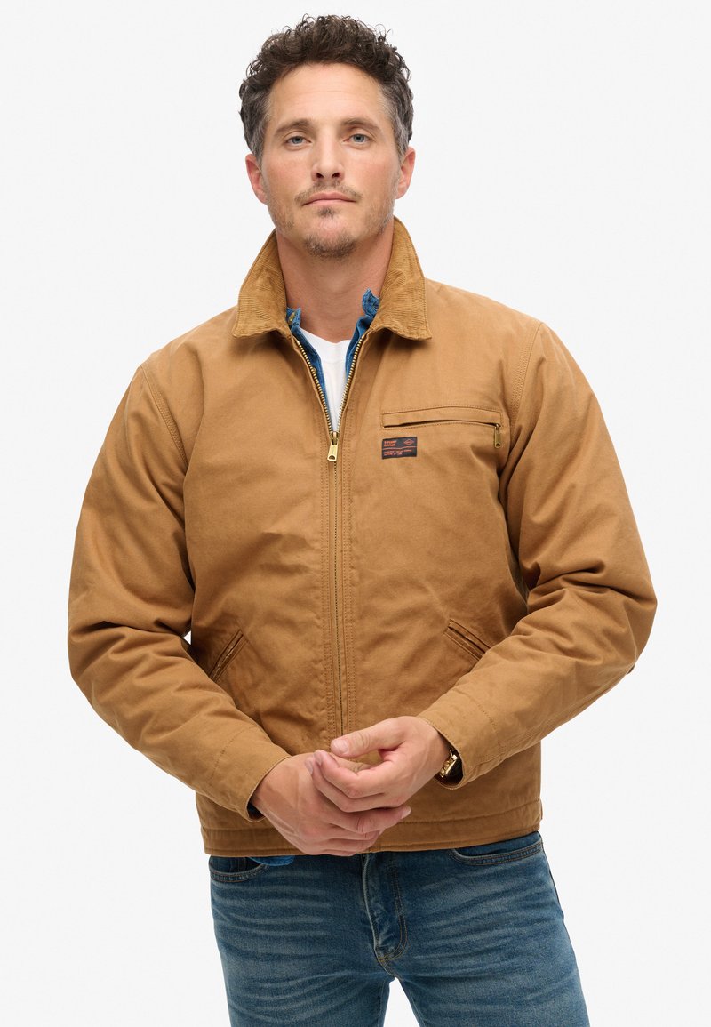 Bruine, ritsvormige jas met een corduroy kraag, zijzakken en een patch op de borst. Draag met een blauw flanellen shirt en een wit T-shirt.