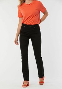 Jean skinny noir associés à un t-shirt orange vif. Le modèle porte des sandales à talons rouges avec une bride texturée. Design et coupe simples.