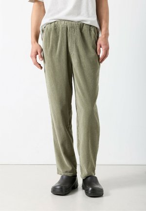 Pantalon en velours côtelé vert olive avec une taille élastique, coupe slim, et tissu texturé. Associé à des chaussures noires. Design simple et minimaliste.