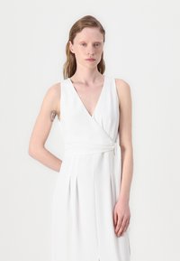 Rochie albă, cu decolteu adânc în V, fără mâneci, fustă plissetată și detalii cu funda pe lateral. Material neted cu un luciu subtil.