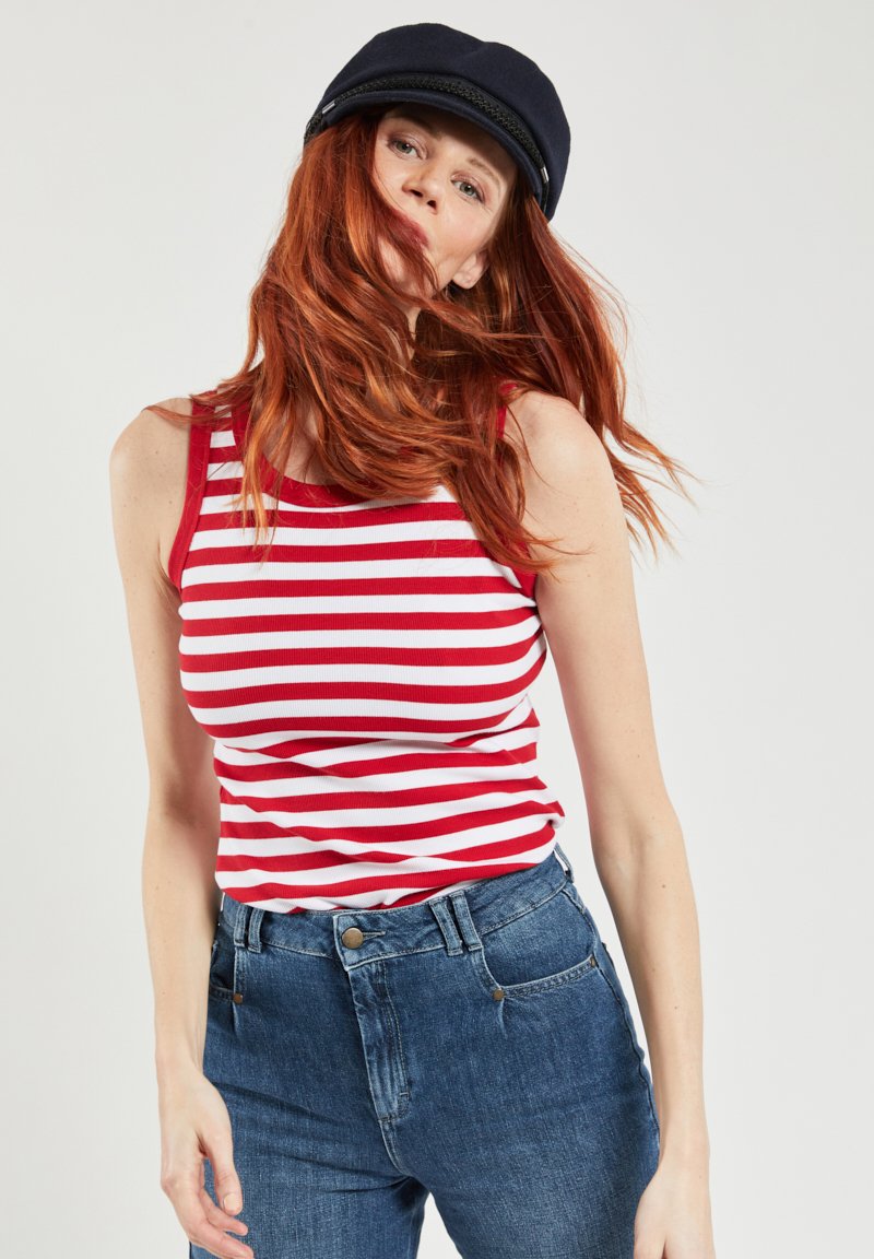 Rood-wit gestreepte tanktop van ribstof, gecombineerd met hoog getailleerde blauwe denim jeans. Model draagt een marineblauwe pet.