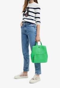 Sac cabas en cuir vert à forme arrondie, poche zippée à l'avant et logo embossé, associé à un pull rayé bleu marine et un jean bleu clair.