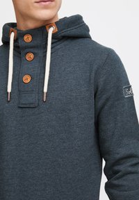Sötét szürke kapucnis jumper, húzózóval, négy fa gombbal és bőrkiegészítővel a galléron. A száron egy kis logós folt található.