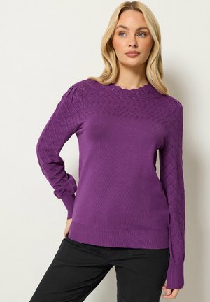 Blonde vrouw met los haar die een paarse gestructureerde gebreide trui en zwarte broek draagt, kijkt naar de zijkant tegen een effen achtergrond.