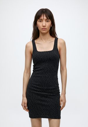 Femme portant une robe noire cintrée sans manches avec de petits pois blancs, debout devant un fond blanc uni.