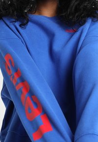 Niebieska bluza z bawełny o mieszanym składzie. Posiada czerwony nadrukowany tekst na lewym rękawie oraz małe czerwone logo na górnej części klatki piersiowej.