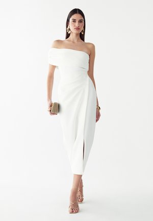 Femme portant une robe midi blanche épaules dénudées avec une fente à l'avant, des talons dorés à brides, de grandes boucles d'oreilles dorées, et tenant une pochette beige.
