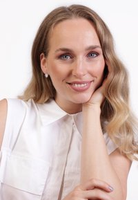 Helles, kragenloses Hemd mit zwei Brusttaschen. Weiche Wellen im langen Haar. Silberne Ohrringe in Herzform. Neutrale Make-up-Töne.