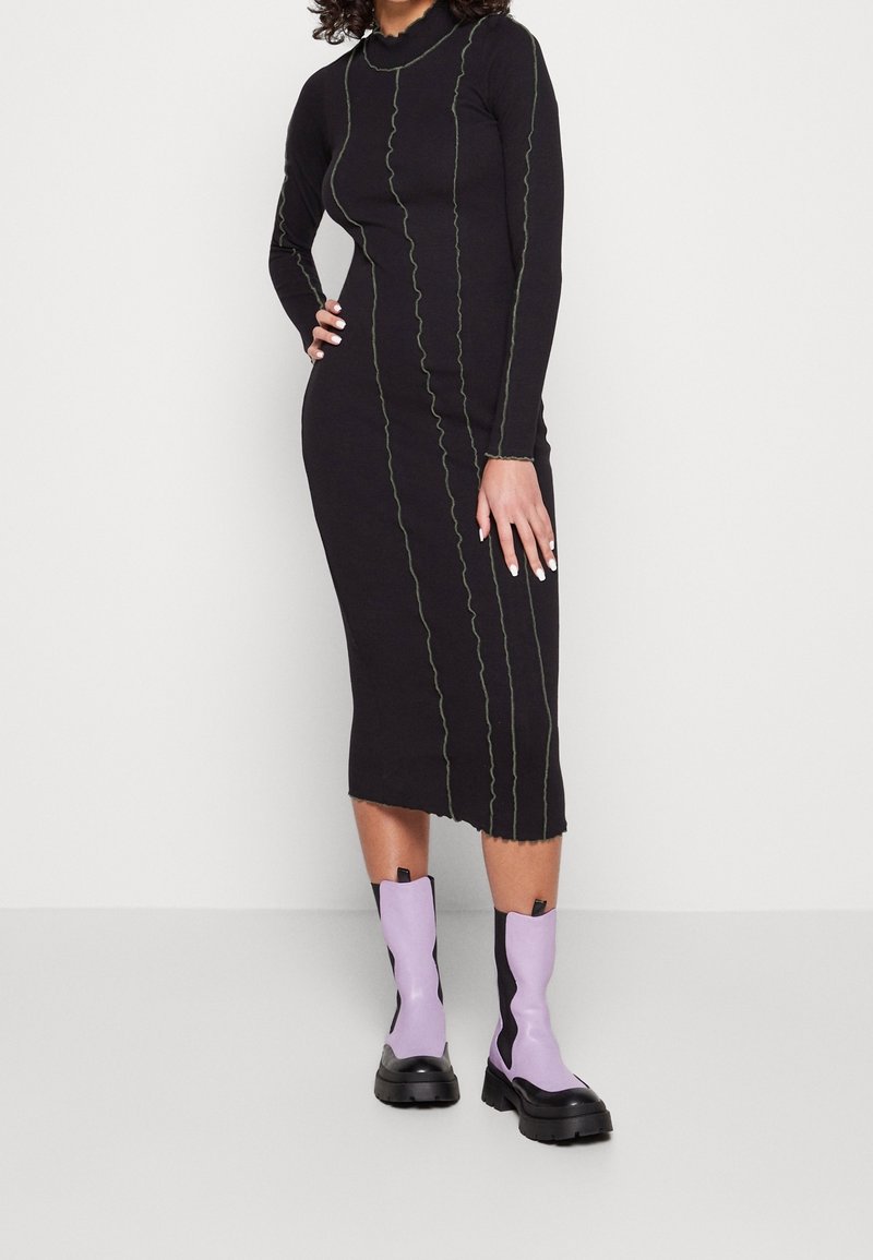 Schwarzes Kleid mit langen Ärmeln und taillierter Form, ausgestattet mit kontrastierenden grünen, wellenförmigen Nähten. Kombiniert mit lilafarbigen Stiefeletten und einer schwarzen Sohle.