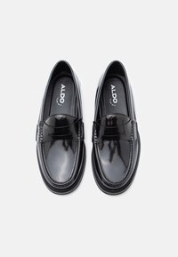 Mocassins en cuir verni noir avec une finition lisse, un bout rond et des accents décoratifs tissés sur le dessus. Logo de la marque visible à l'intérieur.
