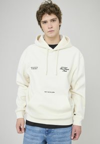 Hoodie crème en tissu doux, doté d'une poche kangourou, de cordons de serrage et d'un texte brodé en noir. Porté avec un jean bleu.