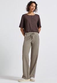 Vrouw staat met haar handen achter haar rug, gekleed in een donkerbruin shirt met korte mouwen, beige joggingsbroek met zijstrepen en witte sneakers.