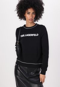 Melna sporta krekls ar baltu "KARL LAGERFELD" logo, ar apaļu kaklu un baltu apdari ap apkakli un malām. Kombinēts ar melnu ādas svārku.