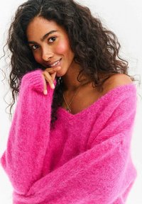 Rozmyty różowy sweter z dekoltem w serek i oversize'owymi rękawami, noszony przez osobę o kręconych włosach, która przykłada palec do ust.