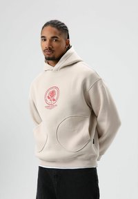 Beige hoodie met grote ronde zakken aan de voorkant, voorzien van een rood grafisch ontwerp van een roos en tekst op de borst. Zachte textuur.