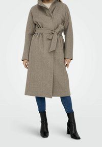 Béžový kabát z vlněné směsi s páskem v pase, dlouhými rukávy a sklápěcím límcem. Nosený přes modré džíny a černé kotníkové boty.
