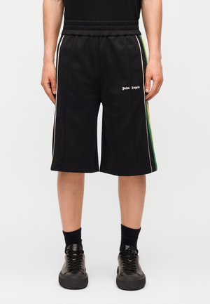 Mężczyzna w czarnych szortach Palm Angels z zielonymi i żółtymi paskami po bokach, czarnych skarpetkach i czarnych trampkach, stojący z rękami wzdłuż ciała.