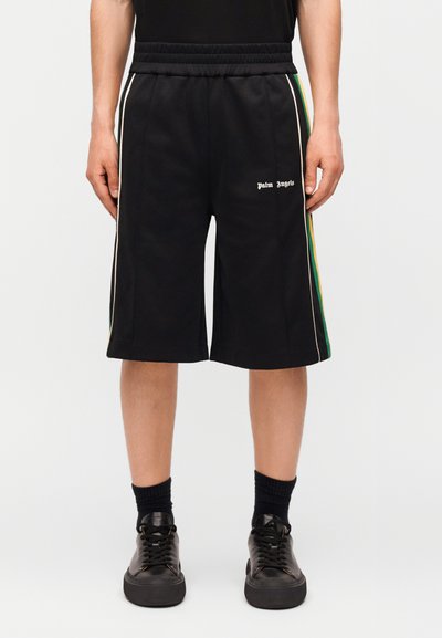 Homme portant un short noir Palm Angels avec des bandes latérales vertes et jaunes, des chaussettes noires et des baskets noires, debout les mains le long du corps.