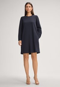 Navyblauwe jurk van gladde stof. Lange mouwen, ronde halslijn en zijzakken. Gestyled met gouden enkelbandhakken.