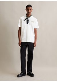 Man in een wit T-shirt met korte mouwen, zwarte broek, zwarte loafers en een gestreepte sjaal om zijn nek, staande tegen een effen beige achtergrond.