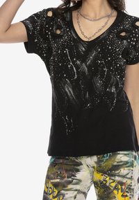 Camiseta negra de cuello en V con agujeros desgastados, acentos metálicos y detalles de pintura blanca salpicada, combinada con pantalones de patrón multicolor.