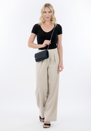 Donna con capelli biondi che indossa una maglietta nera a maniche corte, pantaloni beige a gamba larga, sandali neri, che tiene una piccola borsa nera con tracolla a catena.