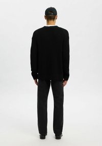 Vyras, apsirengęs juoda megzta megštine, juodomis kelnėmis, juoda kepure ir juodais batais, stovi atsukęs nugarą priešais vientisą baltą foną.