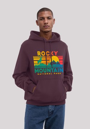 Young man wearing a maroon hoodie with a colorful Rocky Mountain National Park graphic and blue jeans, standing with hands in pockets.