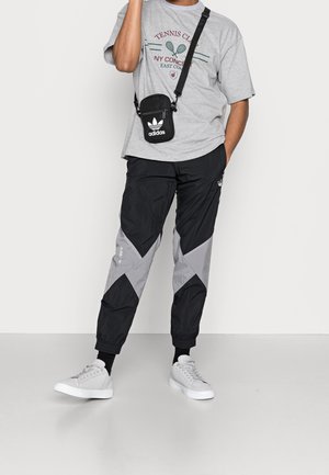 Person trägt ein graues T-Shirt eines Tennisclubs, schwarze und graue Adidas-Trainingshose, weiße Sneakers und eine schwarze Adidas-Umhängetasche.