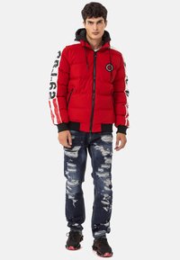Red padded jacket with black accents, zip closure, logo patch, and white striped sleeve details. Paired with ripped dark jeans and black sneakers.