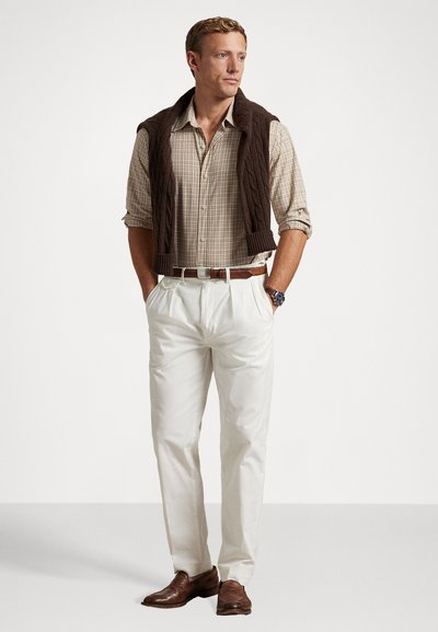 Chemise à carreaux claire sous un gilet en tricot à torsades marron, accompagnée de pantalons plissés blancs et de chaussures en cuir marron. Montre en argent comme accent.