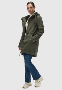 Parka verde oliva con capucha, que presenta un cuello texturizado, bolsillos frontales y un cierre de cremallera con botones. Combinado con jeans acampanados azules y zapatillas beige.