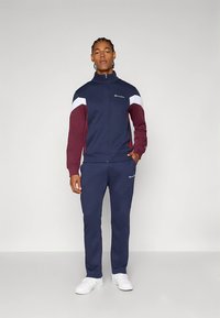 Tengerészkék és bordó melegítő, fehér részletekkel, cipzáras kabát, elasztikus mandzsettákkal és szegéllyel, hozzáillő nadrág, a mellkason és a lábon logóval.