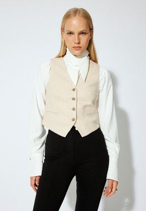 Beige vest med striber og fem knapper, båret over en hvid skjorte med sløjfe-krave. Sammen med sorte tætsiddende bukser for et skræddersyet look.