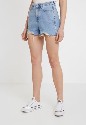 Person iført højtaljede, lyseblå slidte denimshorts og hvide lave sneakers, stående mod en ensfarvet lys baggrund.
