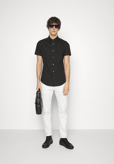 Chemise noire à manches courtes avec boutons, pantalon blanc ajusté, bottes noires à lacets, tenant un sac en cuir noir, portant des lunettes de soleil teintées.