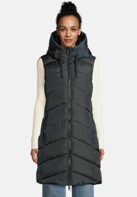 Een vrouw in een mouwloze zwarte puffer vest met capuchon en rits aan de voorkant, over een crèmekleurig geribbeld langemouwen top en blauwe jeans.