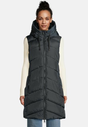 Een vrouw in een mouwloze zwarte puffer vest met capuchon en rits aan de voorkant, over een crèmekleurig geribbeld langemouwen top en blauwe jeans.