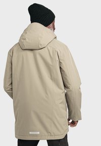 Beige wasserdichte Jacke mit Kapuze, strukturiertem Stoff und geradem Schnitt. Mit einem Logo-Akzent am Saum auf der Rückseite. Eine schwarze Mütze wird getragen.