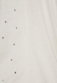Hellbeige Strickstoff mit einer vertikalen Reihe von kleinen, glänzenden Strasssteinen an einer Seite, die einen dekorativen Akzent zur Textur hinzufügen.