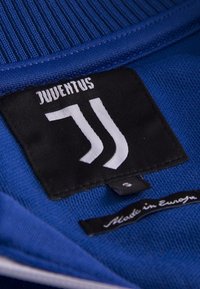 Blå klädesetikett med "JUVENTUS" i vit text på en svart bakgrund, med "JJ"-logotypen och en etikett "Tillverkad i Europa" nedan.