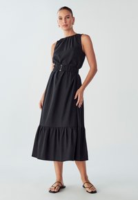 Donna che indossa un vestito midi senza maniche di colore nero con una cintura in vita, orecchini a cerchio dorati e sandali neri flat con cinturini, in piedi su uno sfondo neutro.