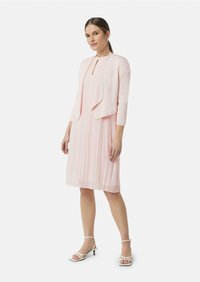 Rosa plissiertes Kleid mit rundem Ausschnitt, begleitet von einer passenden kurzen Jacke. Weiße Riemchen-Absätze runden das Outfit ab.