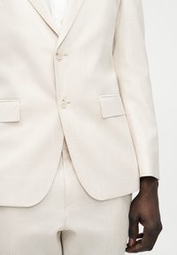 Ανοιχτό μπεζ σακάκι με ταιριαστό παντελόνι σε άτομο με σκούρο δέρμα, εμφανίζοντας κουμπιά, τσέπες με καπάκι και χαλαρό χέρι στα πλάγια.