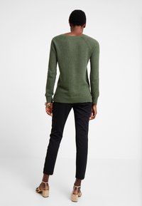 Maglione verde a trama con maniche lunghe e scollatura rotonda, abbinato a pantaloni slim-fit neri e sandali con tacco beige.